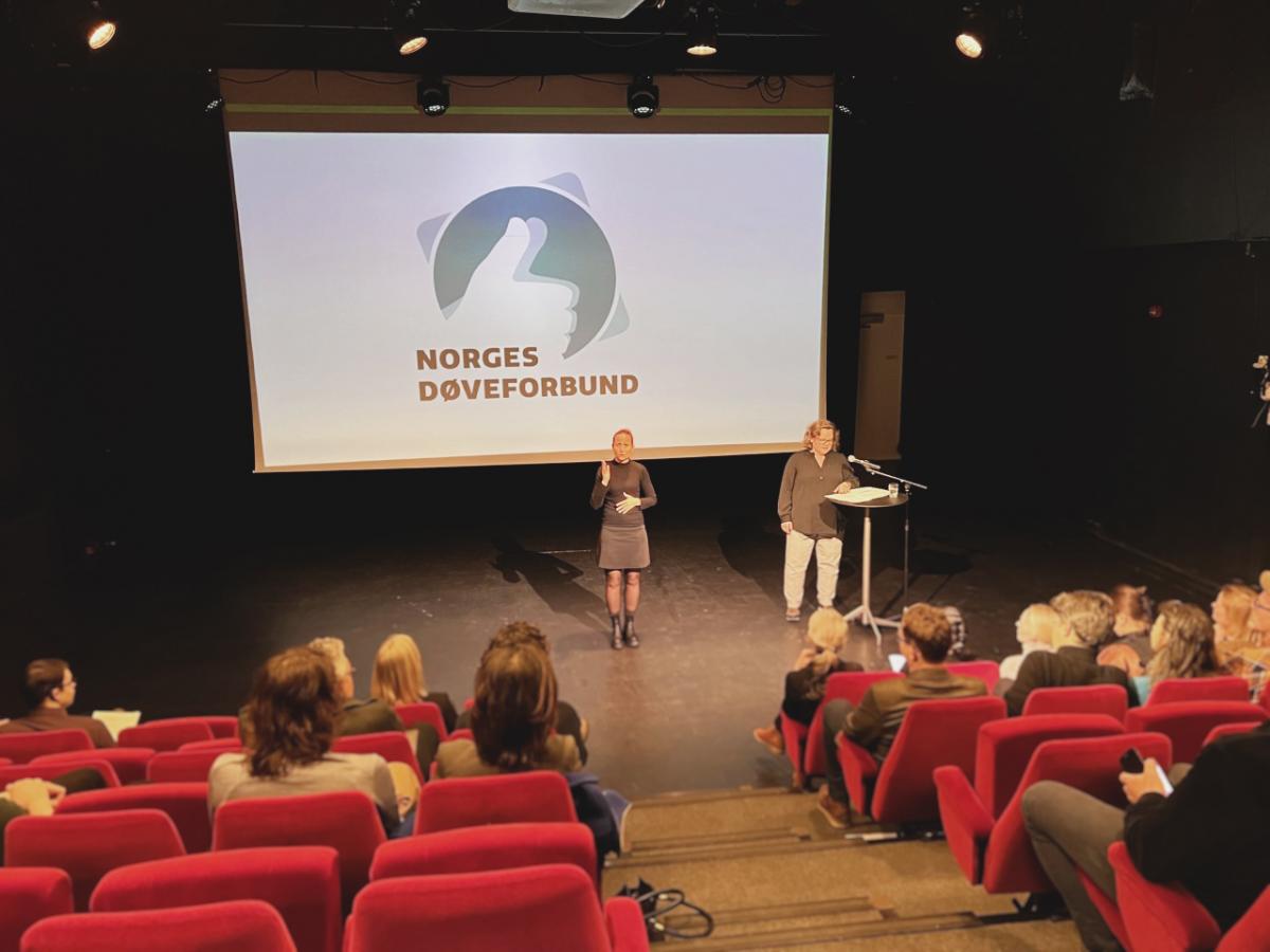 Elisabeth Frantzen Holte holder et foredrag. I midten står en tegnspråktolk som oversetter fra tale til tegnspråk.