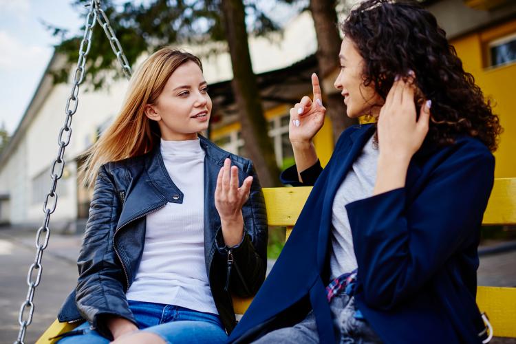 To damer sitter ute, vendt mot hverandre og prater sammen på tegnspråk. 